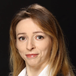 Professional headshot: woman with light brown hair in a white collared shirt against a dark background, neutral expression.